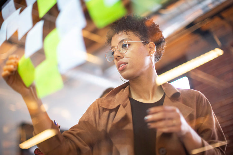 A person wearing glasses and a brown jacket arranges sticky notes on a glass wall, focusing intently on their task in a modern, well-lit workspace.