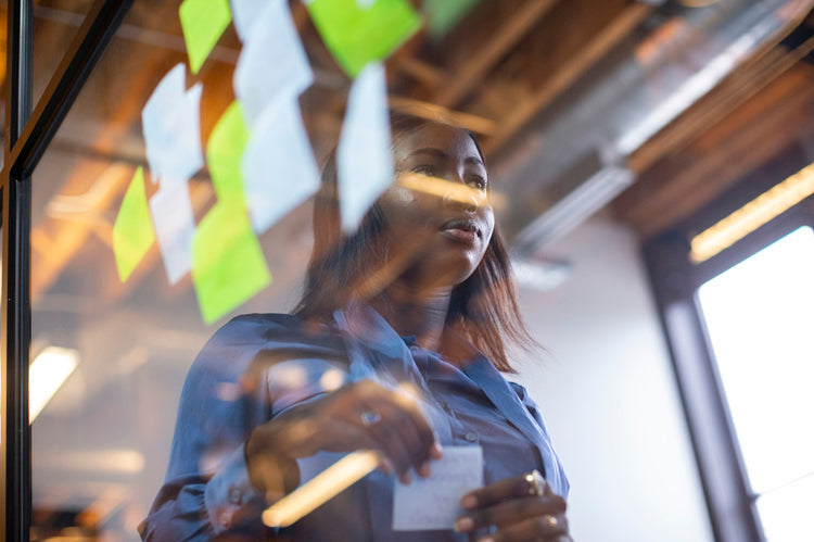 A woman stands behind a glass wall covered with colorful sticky notes, holding a note in her hand, appearing focused and thoughtful in a modern office setting with exposed beams and bright lighting.