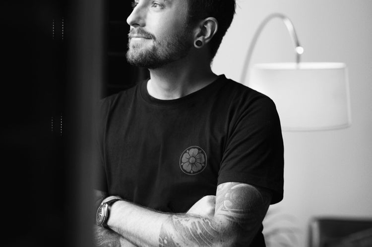 A man with tattoos and a beard stands by a window, looking outside with his arms crossed. He wears a black t-shirt and has stretched earlobes. The scene is in black and white, with a lamp and chair in the background.