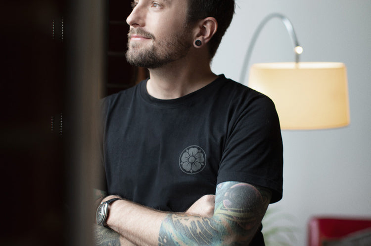 A bearded man with tattoos and stretched earlobes stands indoors, arms crossed, looking thoughtfully out a window. He wears a black T-shirt. A lamp and a red couch are visible in the background.