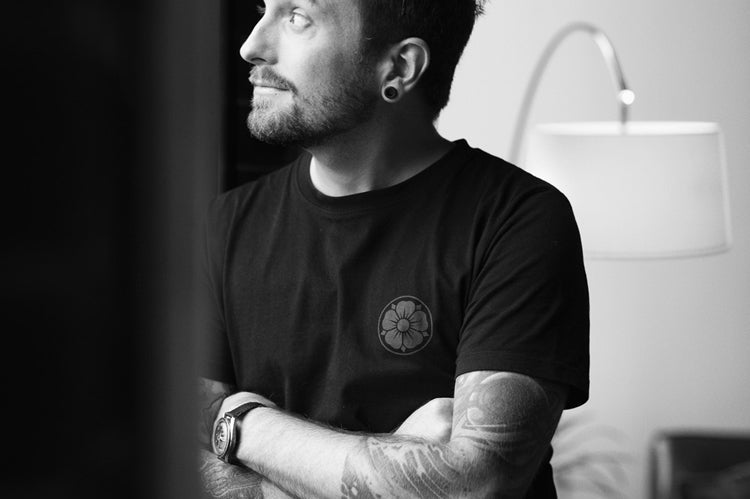 A man with tattoos and a beard, wearing a black t-shirt, stands indoors with his arms crossed, looking out of a window. There is a lamp and a sofa in the background. The image is in black and white.