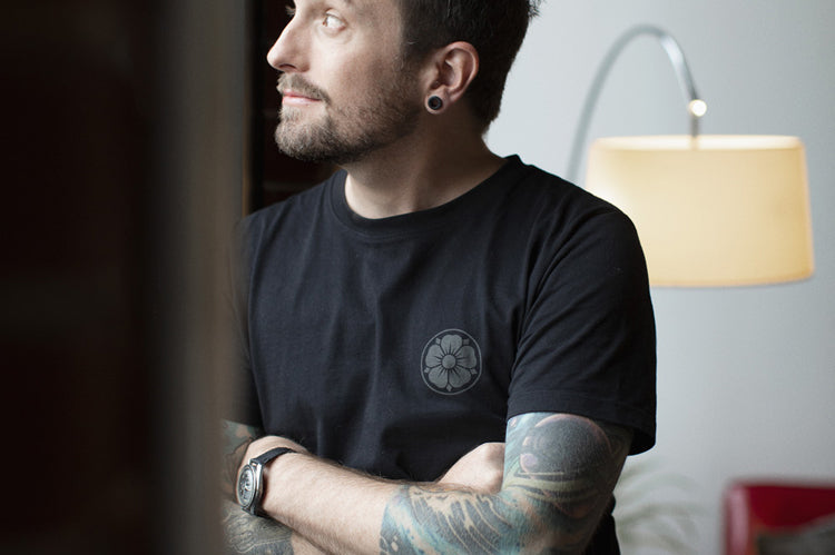 A man with tattoos and a beard, wearing a black t-shirt, stands by a window with his arms crossed, looking outside and smiling. A floor lamp and red chair are visible in the modern room behind him.
