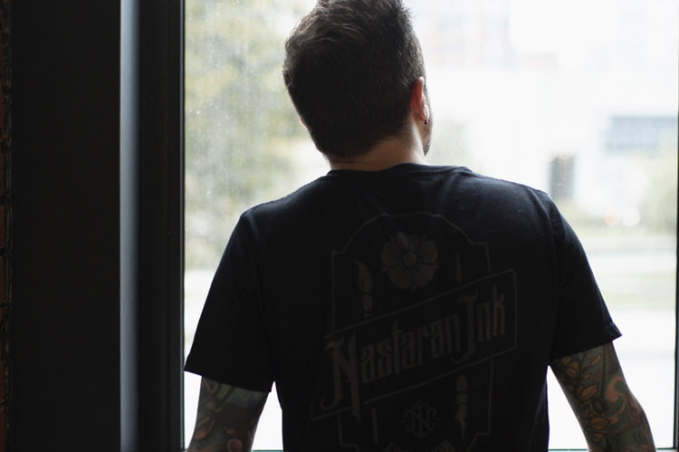 A person with tattooed arms, wearing a black Nastaran Ink t-shirt, stands indoors facing a large window with both hands resting on the windowsill, looking outside.