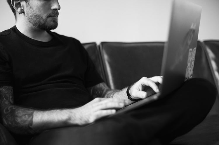 A man with tattoos sits on a couch, wearing a black t-shirt, and types on a laptop resting on his lap. The image is in black and white and only shows the lower half of his face and upper body.