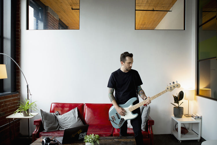 A man in a black t-shirt plays a light blue electric bass guitar while sitting on a red couch in a modern, minimalist living room with a laptop and plants.