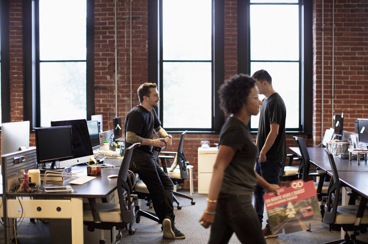 Three people in a modern office with brick walls and large windows; one person walks by holding papers, while two others stand and sit near desks with computers, engaged in conversation.