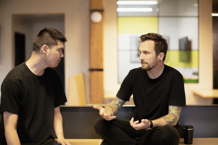 Two men sitting indoors, engaged in a serious conversation. Both wear black t-shirts; one gestures with his hands while the other listens intently. The background shows a modern office setting with wooden beams and glass walls.