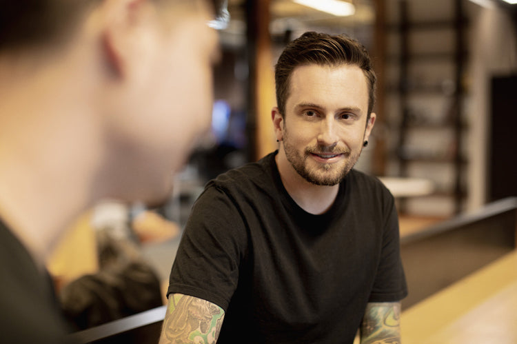A man with tattoos and a trimmed beard, wearing a black t-shirt, smiles and listens to another person in a blurred foreground. They appear to be having a conversation in a modern indoor setting.