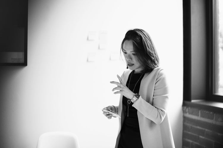 A woman in business attire stands indoors near a window, holding a card and gesturing with her hand as she speaks, appearing focused and engaged in conversation.