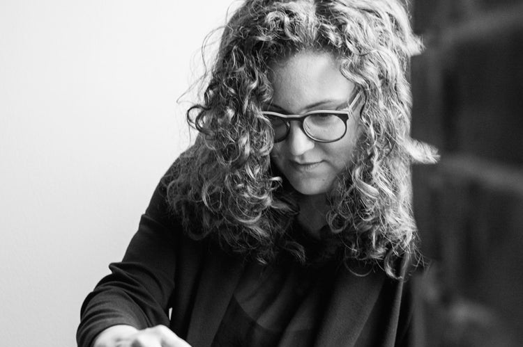 Black and white photo of a person with curly hair and glasses, wearing a dark top, sitting and using a tablet or laptop, with part of a brick wall visible in the foreground.