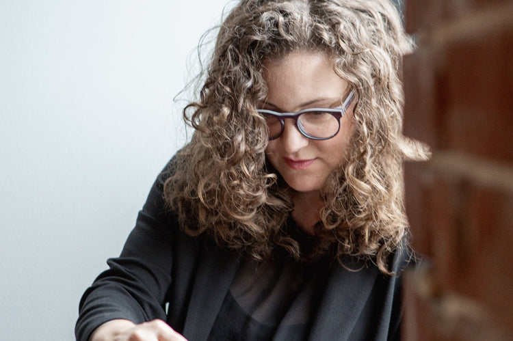 A woman with curly hair and glasses, wearing a black blazer, sits by a brick wall and looks down at a laptop or tablet in her hands.