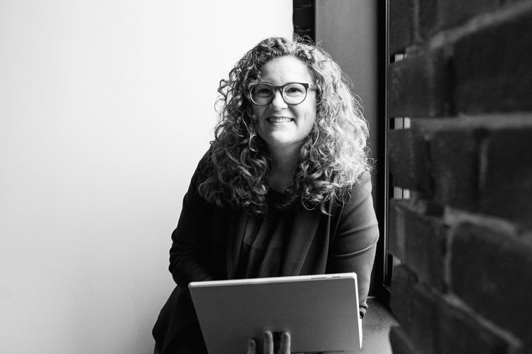 A smiling person with curly hair and glasses stands by a window holding an open laptop, dressed in a blazer and jeans. The photo is black and white with a brick wall in the foreground.