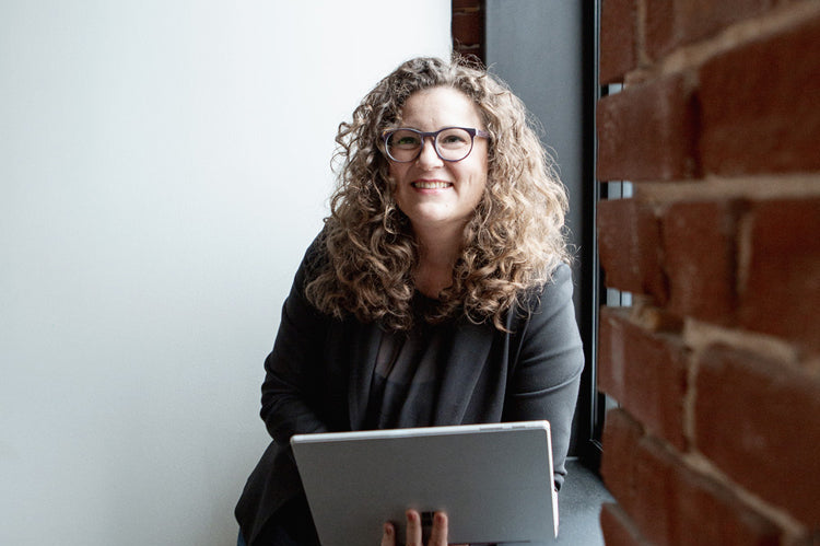 A woman with curly hair and glasses, wearing a dark blazer and jeans, smiles while holding a laptop and leaning against a windowsill next to a brick wall.