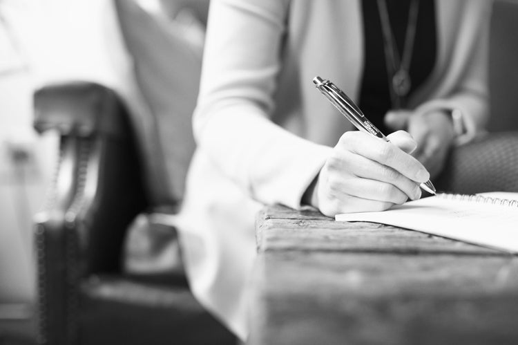 A person in a blazer writes with a pen on paper at a rustic wooden table. The image is in black and white and focuses on the hand and pen, with the background softly blurred.