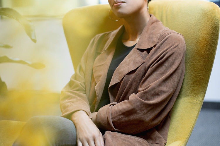 A person with short curly hair sits in a yellow armchair, wearing a brown coat, black shirt, and jeans, looking thoughtfully at the camera in a modern indoor setting.