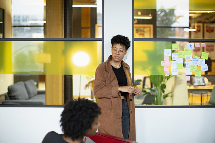 A person in a brown coat stands by a glass wall covered with colorful sticky notes, speaking to another person seated in the foreground. The setting appears to be a modern office with large windows.