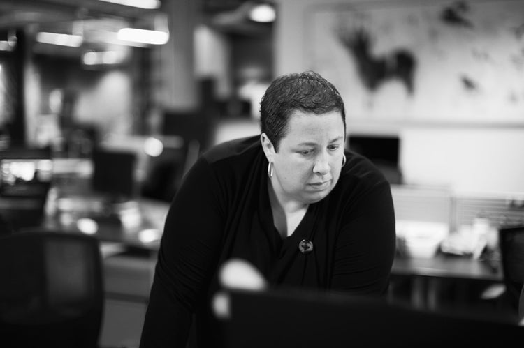 A person with short hair and a black top leans forward, looking intently at something off-camera, in a modern, blurred office setting.