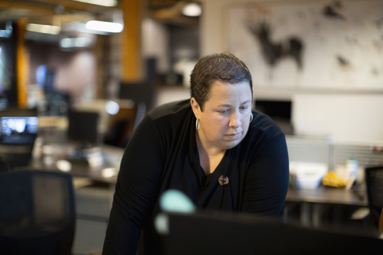 A person with short hair and a black top leans over a desk, looking intently at a computer screen in a modern office setting with blurred background.