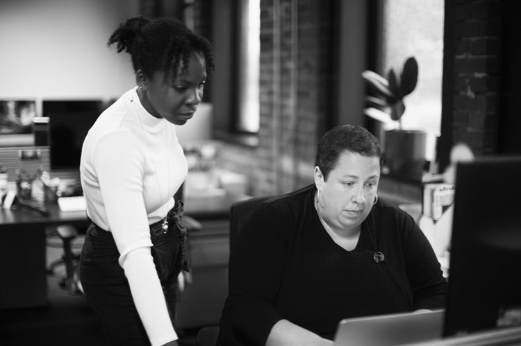 Two women in an office; one is seated and working on a computer while the other stands beside her, looking at the screen. The setting is modern with brick walls and large windows.