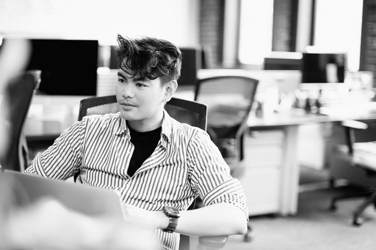 A person with short, wavy hair sits at a desk in a modern office, looking to the side. They wear a striped shirt over a dark top. The background shows empty chairs, desks, and computers. The image is in black and white.