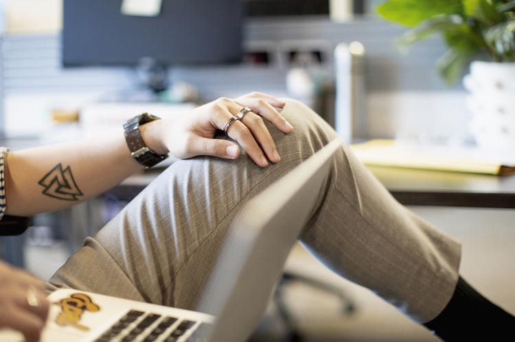 A person wearing light gray pants rests their hand on their crossed knee while working on a laptop. The individual has a geometric tattoo, wears rings, and sits in a modern office setting with plants in the background.