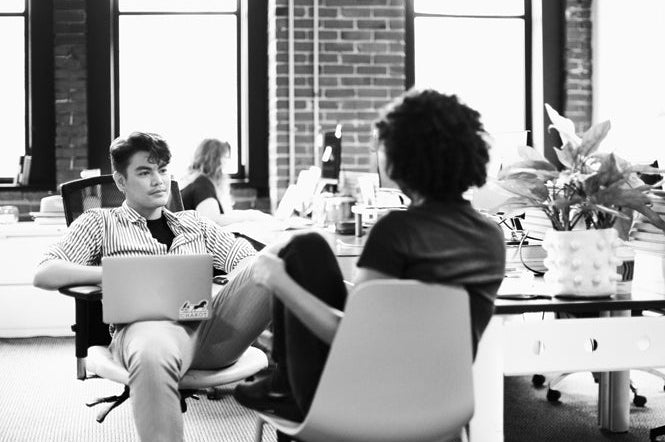 Two people sit facing each other in a modern office with large windows and brick walls. One has a laptop on their lap. A plant and other desks are visible in the background. The image is black and white.