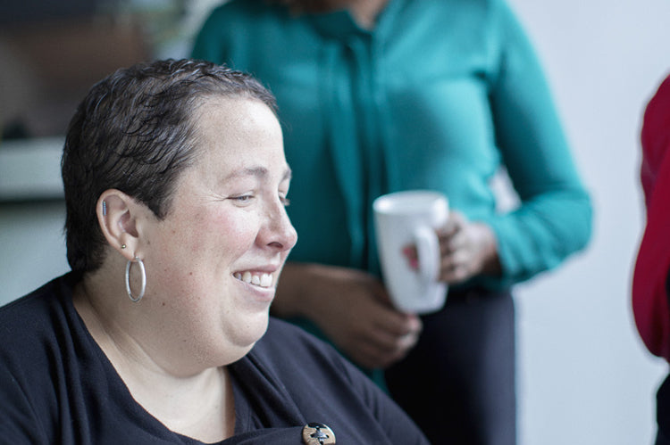 Two women in an office setting; one is seated and smiling while working, and the other stands in the background holding a mug, both appearing engaged and positive.