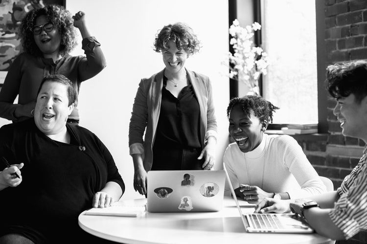 A group of five people sit and stand around a table with laptops, laughing and smiling together in a brightly lit office space.