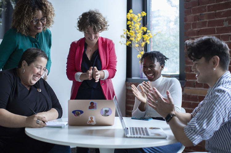 Five people gather around a table with laptops, laughing and clapping together in a bright room with exposed brick walls and a vase of yellow flowers, suggesting a joyful team meeting or collaboration.