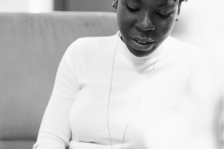 A woman sits on a couch, looking down and using a tablet. She wears a white turtleneck sweater, a necklace, and dark pants. The setting appears to be indoors, possibly an office or lounge. The photo is in black and white.