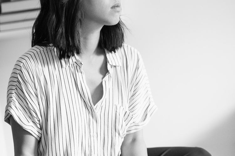 A person with shoulder-length dark hair, wearing a striped shirt, sits indoors looking thoughtfully to the side. Papers and a pen are in their hands and lap. The background includes shelves with books and a decorative hand sculpture.