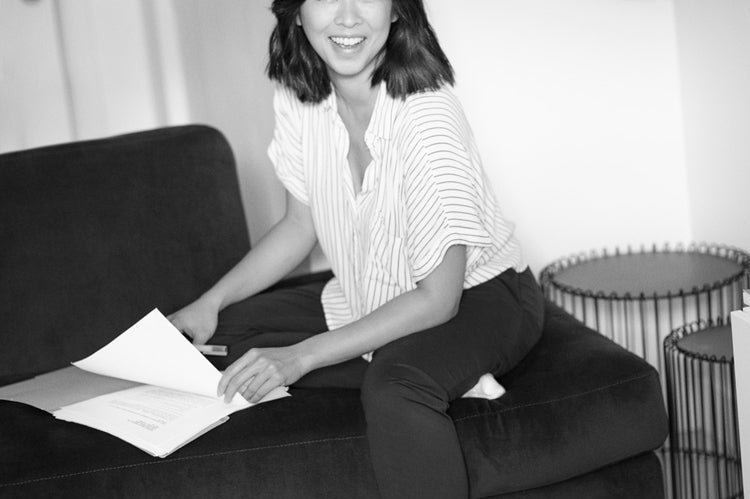 A woman sits on a dark couch, smiling and holding papers. She wears a striped shirt, dark pants, and one sock. The room has a cozy feel, with shelves, a wire basket, sandals on the floor, and light streaming in. Black and white photo.