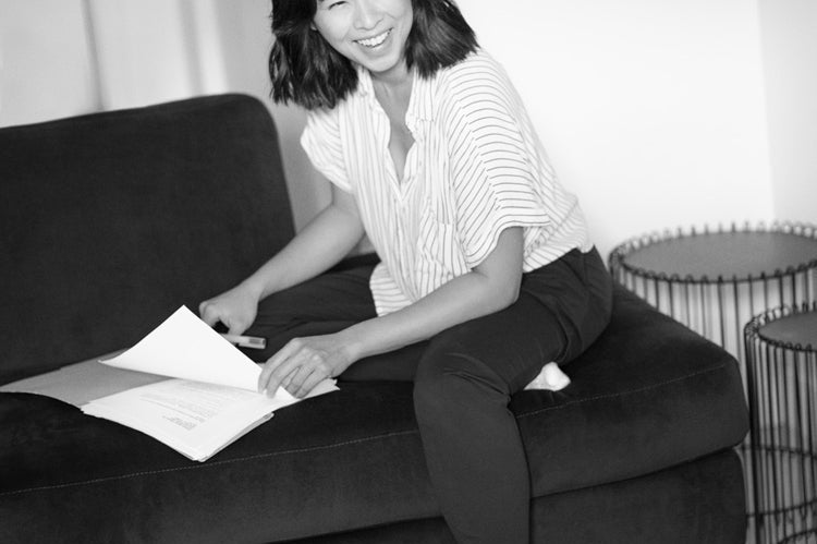 A woman sits cross-legged on a sofa, smiling while holding papers. She wears a striped shirt, dark pants, and white socks. Sandals are on the floor beside her on a cowhide rug in a cozy, modern room.