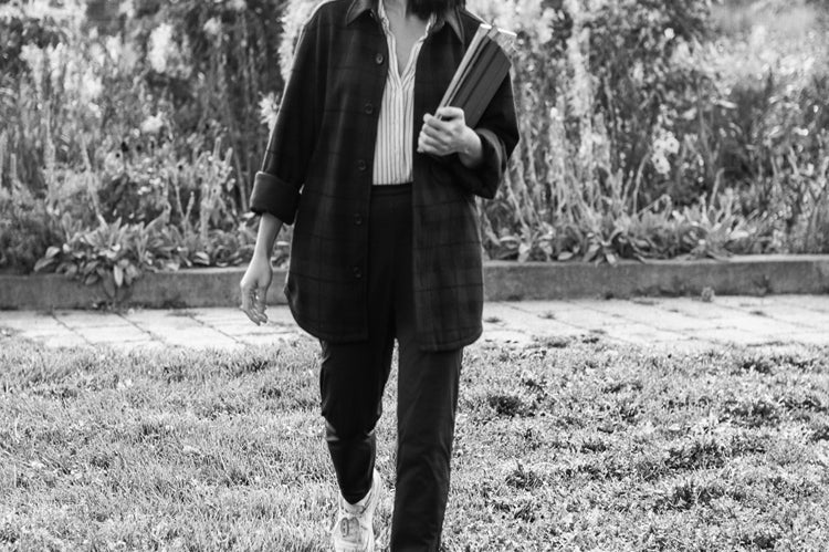 A person walks on grass, holding a notebook or folder, and looks to the side. They wear a long-sleeve shirt, pants, and sneakers. Blurred greenery and buildings are visible in the background. Black and white photo.
