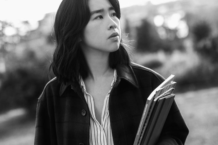 A person with shoulder-length dark hair holds books and notebooks while standing outdoors. They wear a striped shirt and a dark jacket, looking thoughtfully to the side. The background is blurred greenery. The photo is black and white.