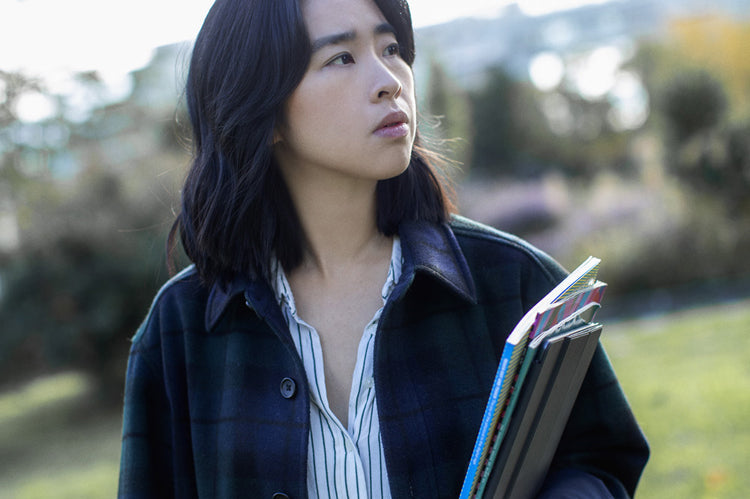 A person with shoulder-length dark hair, wearing a plaid coat over a striped shirt, stands outdoors holding notebooks and folders, looking thoughtfully to the side. The background is grassy with blurred trees.