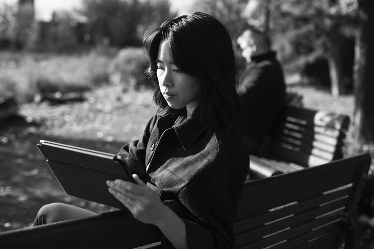A woman sits on a park bench reading a tablet, with sunlight on her face. In the background, a man sits facing away on the same bench. The image is in black and white.