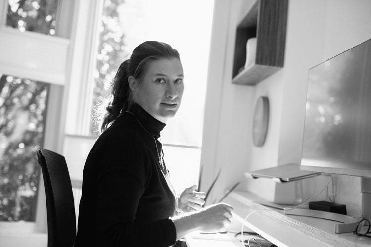 A woman in a black turtleneck sits at a desk by a window, turning to look at the camera. Sunlight streams in, and a computer monitor and shelves are visible in the modern workspace.
