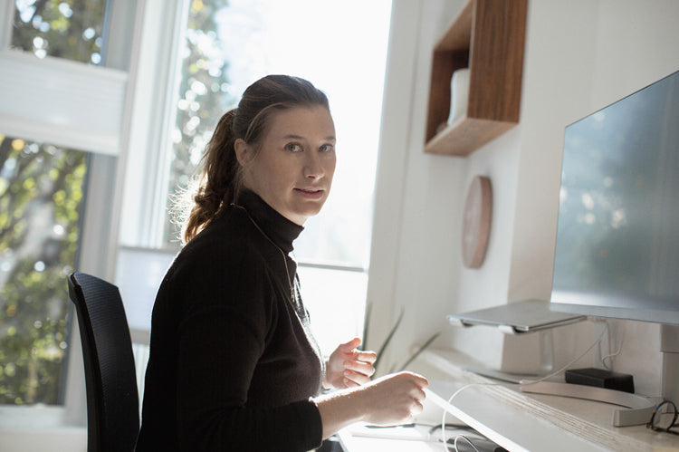 A woman with light brown hair in a ponytail sits at a desk in a bright, modern office, looking towards the camera. She wears a black turtleneck and is near a computer monitor with sunlight streaming in.