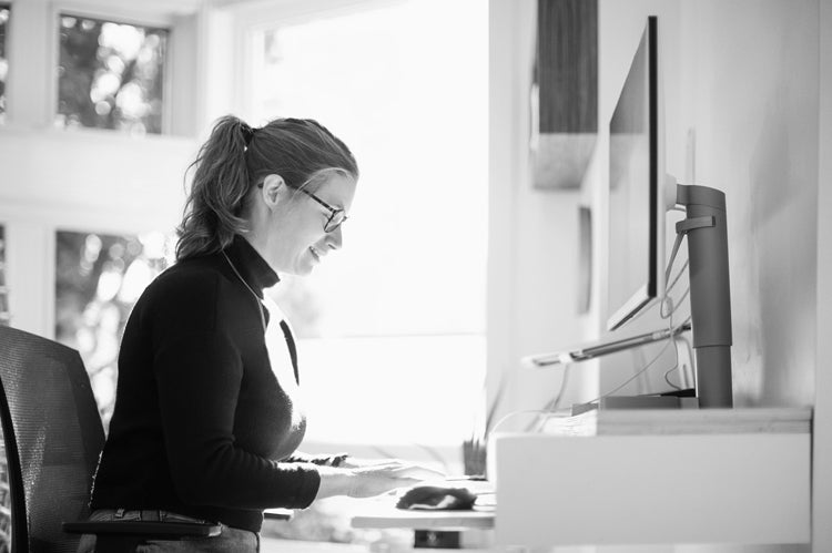 A woman wearing glasses and a turtleneck sweater sits at a desk, typing on a keyboard in front of a computer monitor, with sunlight streaming through large windows in the background.