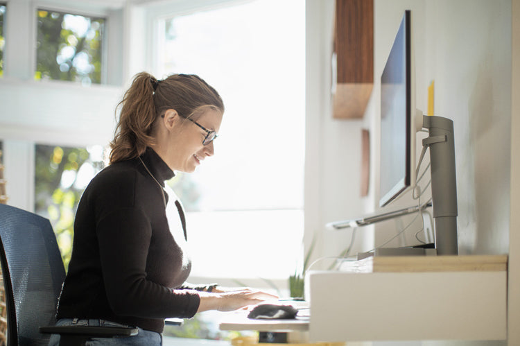 A woman with glasses and a ponytail is sitting at a desk and typing on a keyboard in front of a large computer monitor in a bright, sunlit room.