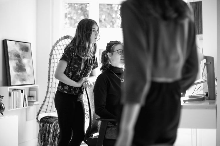 Three people in a home office; one seated at a desk using a computer, another standing beside and looking at the screen, and a third person in the foreground, partially out of focus. The scene is in black and white.