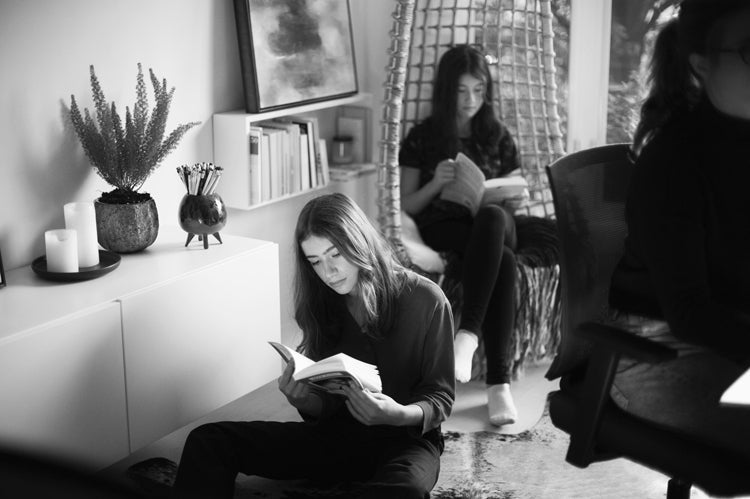 Three young people reading indoors. One sits on the floor near a cabinet with candles and a plant, another sits in a hanging chair by a window, and the third is partially visible, seated at a desk. The image is in black and white.