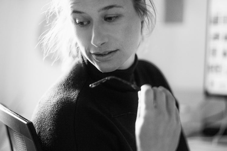 A person with light hair pulled back, wearing a dark top, sits on an office chair and looks to the side while holding a pair of eyeglasses. The background is softly blurred, suggesting an indoor workspace.