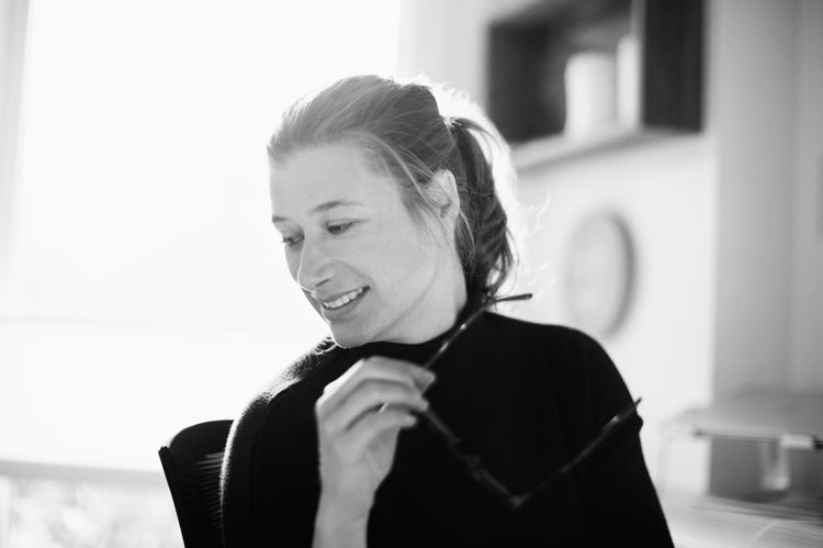 A woman with light hair tied back smiles while looking down, holding eyeglasses in her hand. She is sitting indoors in a softly lit, modern room with blurred shelves in the background.