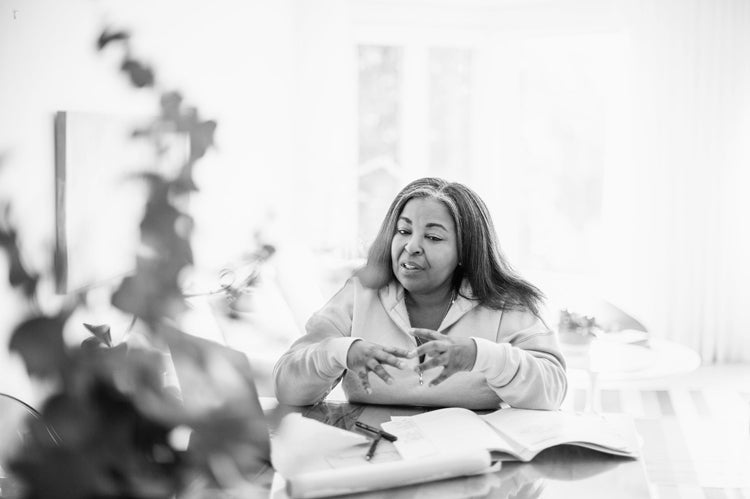 A woman sits at a table with open books and papers, gesturing with her hands as she talks, with natural light streaming in from a window behind her. The image is in black and white.