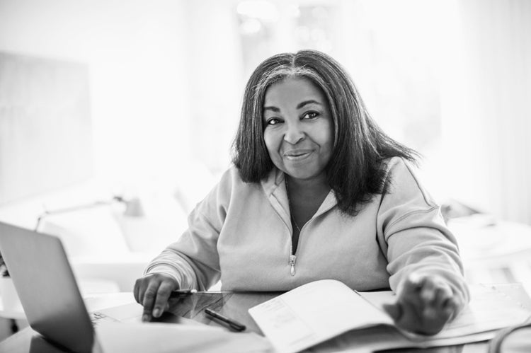A woman with shoulder-length hair sits at a desk, smiling, with a pen in hand and papers in front of her next to an open laptop. The scene is bright and set in a home or office environment.