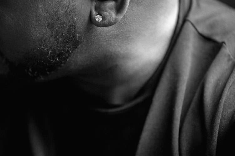 Black and white close-up of a mans head and shoulder, focusing on his ear with a diamond stud earring, trimmed beard, and smooth bald scalp. The man is facing away from the camera, creating a moody, intimate atmosphere.
