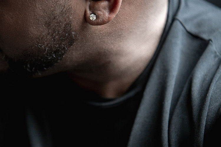 Close-up of a mans ear wearing a diamond stud earring; only part of his face, bald head, and dark shirt are visible, with the background out of focus.
