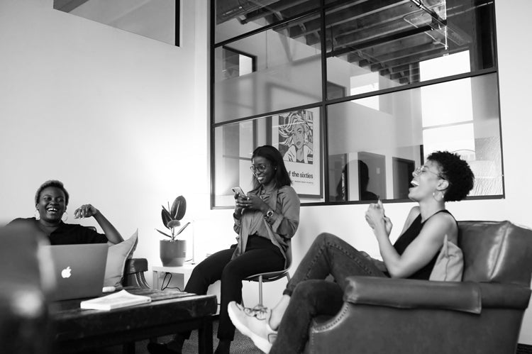 Three people are sitting and laughing together in a modern office space, one with a laptop, another with a phone, and large windows and a potted plant in the background. The atmosphere is relaxed and friendly.